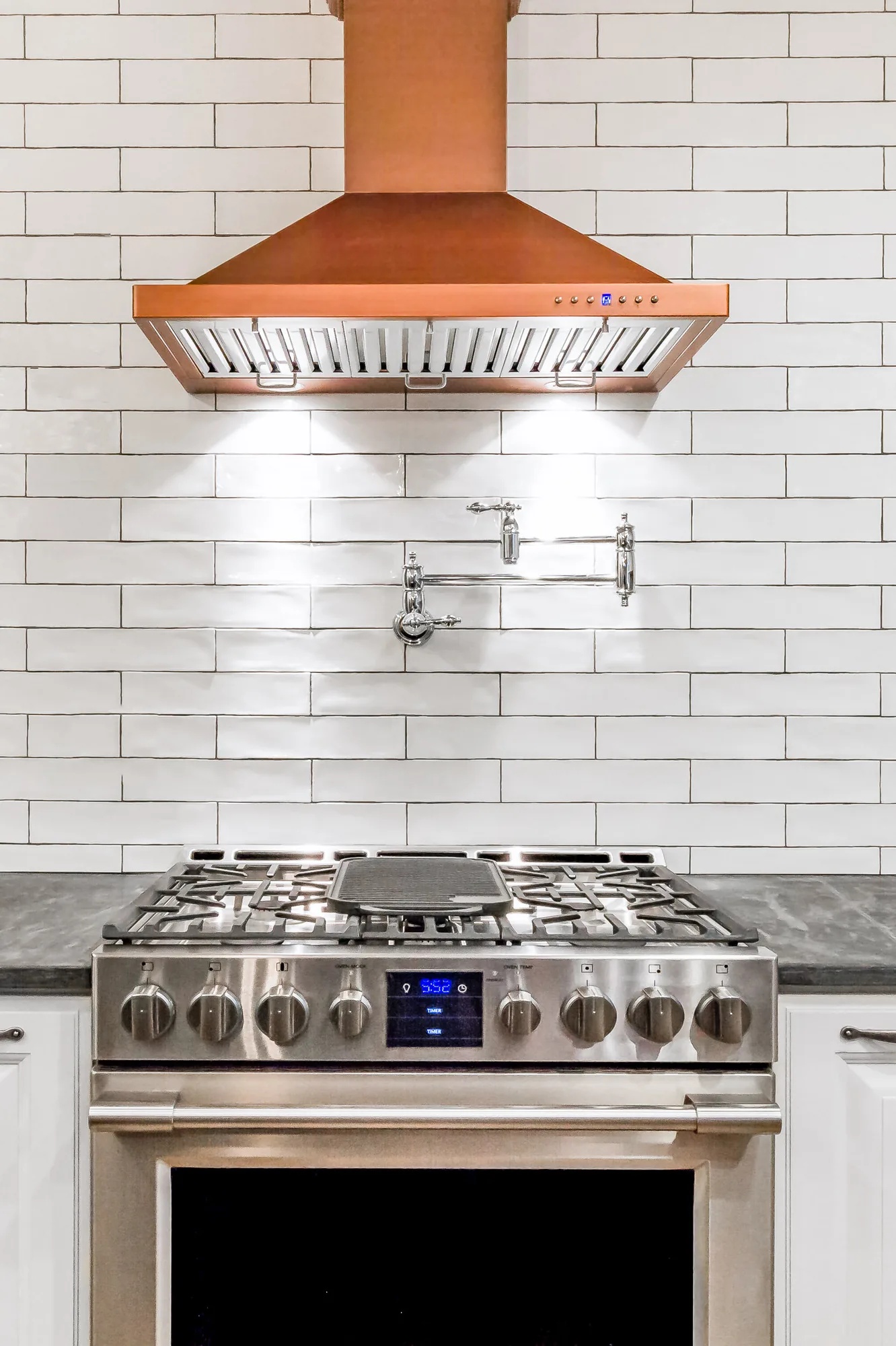 Stainless gas range under a copper range hood and white subway tile
