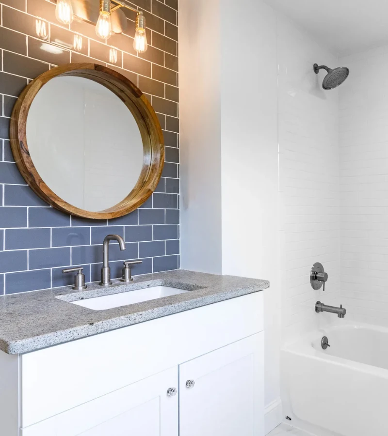 Newly built bathroom with a blue subway-tile wall, round wood-framed mirror, granite vanity, and a tiled tub and shower